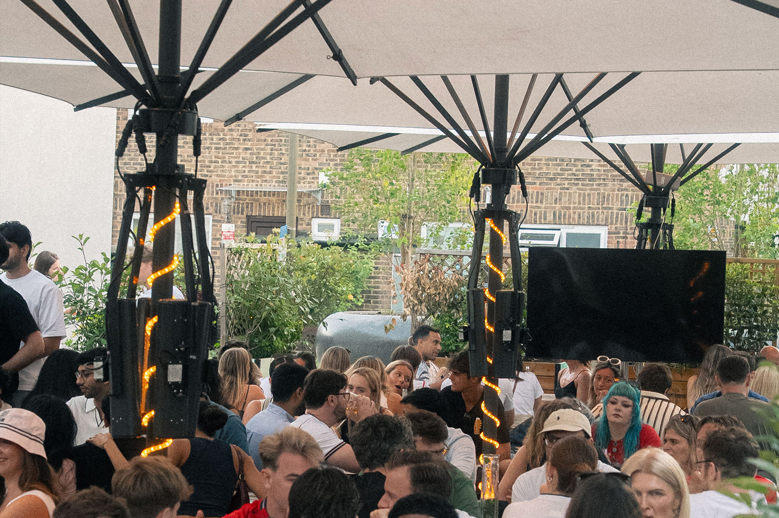 Groups of people sitting outside under umbrellas watching a tv screen in a beer garden.