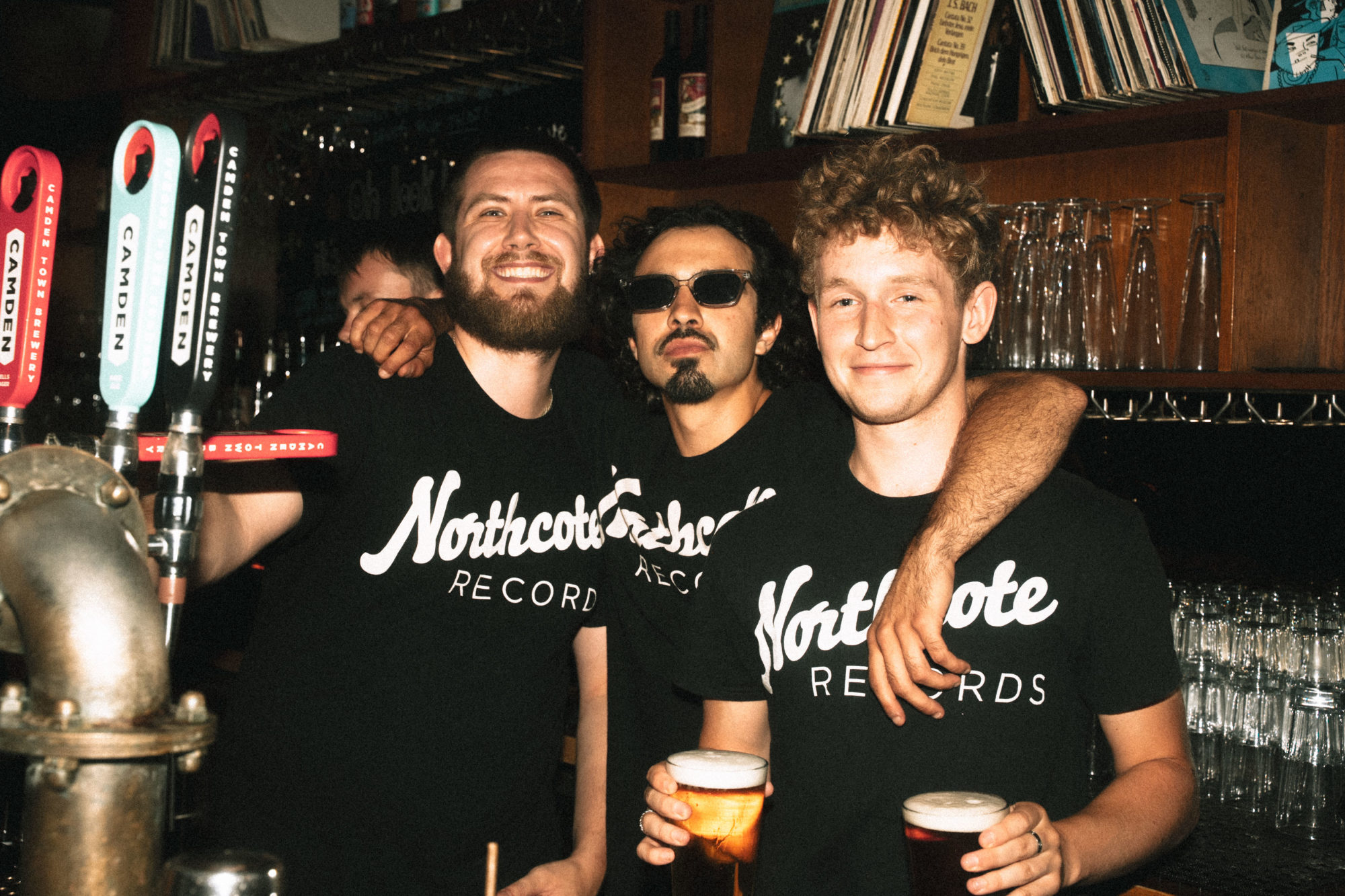 Three male bartenders holding pints of beer with a wall of vinyls behind them.